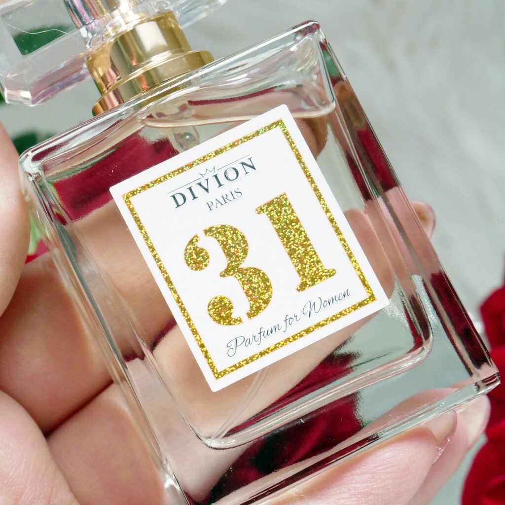 Zbliżenie na etykietę i detale flakonu lanych perfum Divion 31 Zbliżenie na złotą etykietę przezroczystego flakonu lanych perfum Divion 31 trzymanego w dłoni.