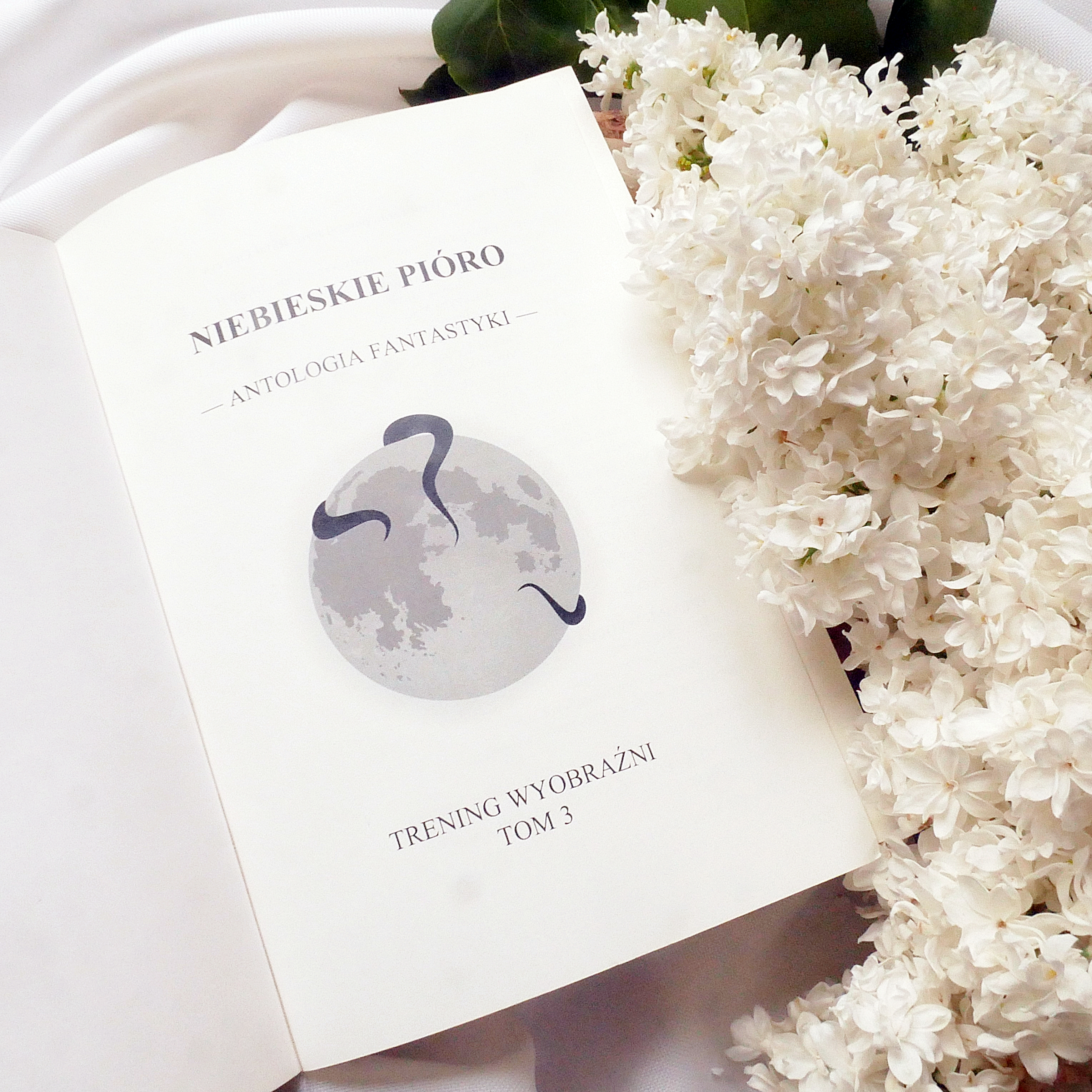Strona tytułowa antologii „Niebieskie Pióro” z grafiką księżyca i białym bzem. Strona tytułowa książki „Niebieskie Pióro. Antologia Fantastyki” leżąca na białym materiale obok gałązek białego bzu.
