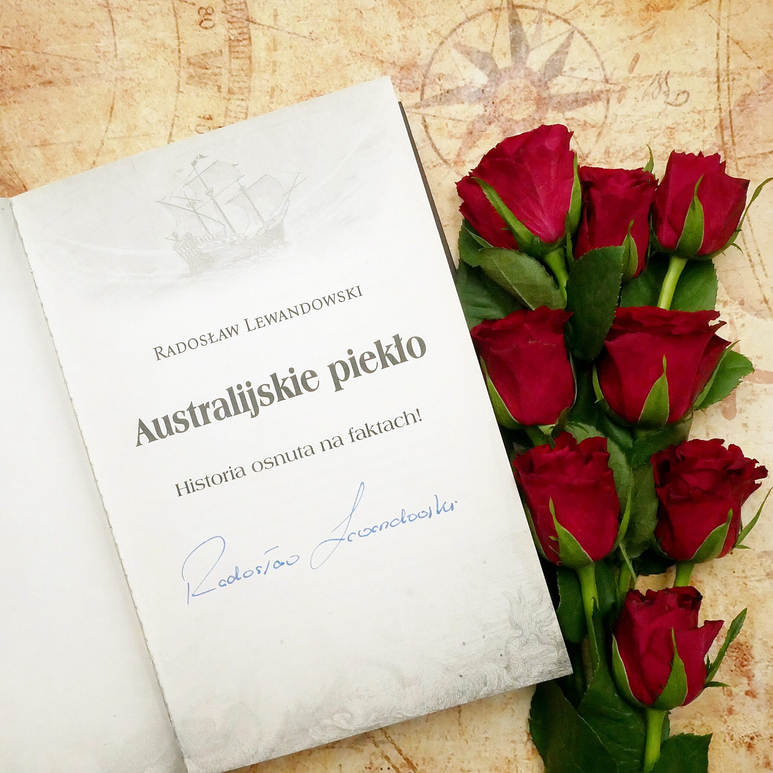 Strona tytułowa książki „Australijskie piekło” z autografem autora Strona tytułowa książki „Australijskie piekło” z podtytułem „Historia osnuta na faktach!” i autografem autora. Książka leży na tle starej mapy obok czerwonych róż.