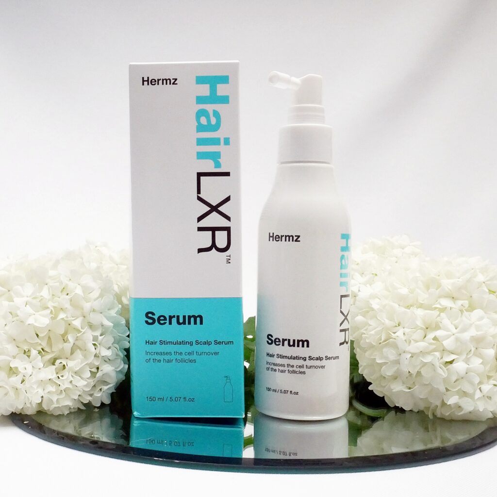 Skoncentrowane serum HairLXR Dermz. Skoncentrowane serum do skóry głowy HairLXR z aplikatorem i logo Hermz (obecna nazwa Dermz) ustawione na lustrzanej tacy z białymi hortensjami.