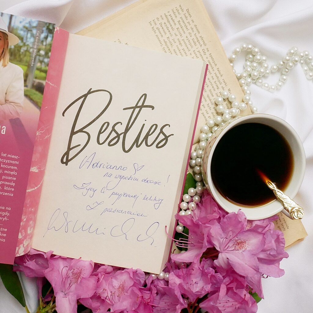 Strona tytułowa książki Magdalena Winnicka „Besties na zawsze” z dedykacją autorki dla Adrianny, autorki bloga nawysokimobcasie.pl, leżąca wśród różowych kwiatów, obok filiżanki kawy i sznura pereł na białym materiale.