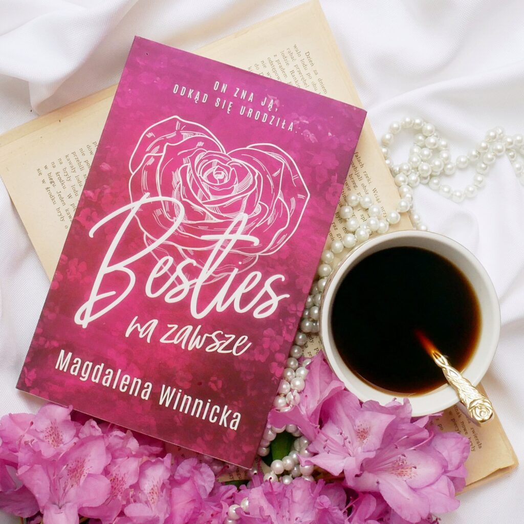 Różowa okładka książki Magdalena Winnicka „Besties na zawsze”” z grafiką białej róży, leżąca na białym materiale i starym papierze, obok filiżanki z kawą, różowych kwiatów i pereł.