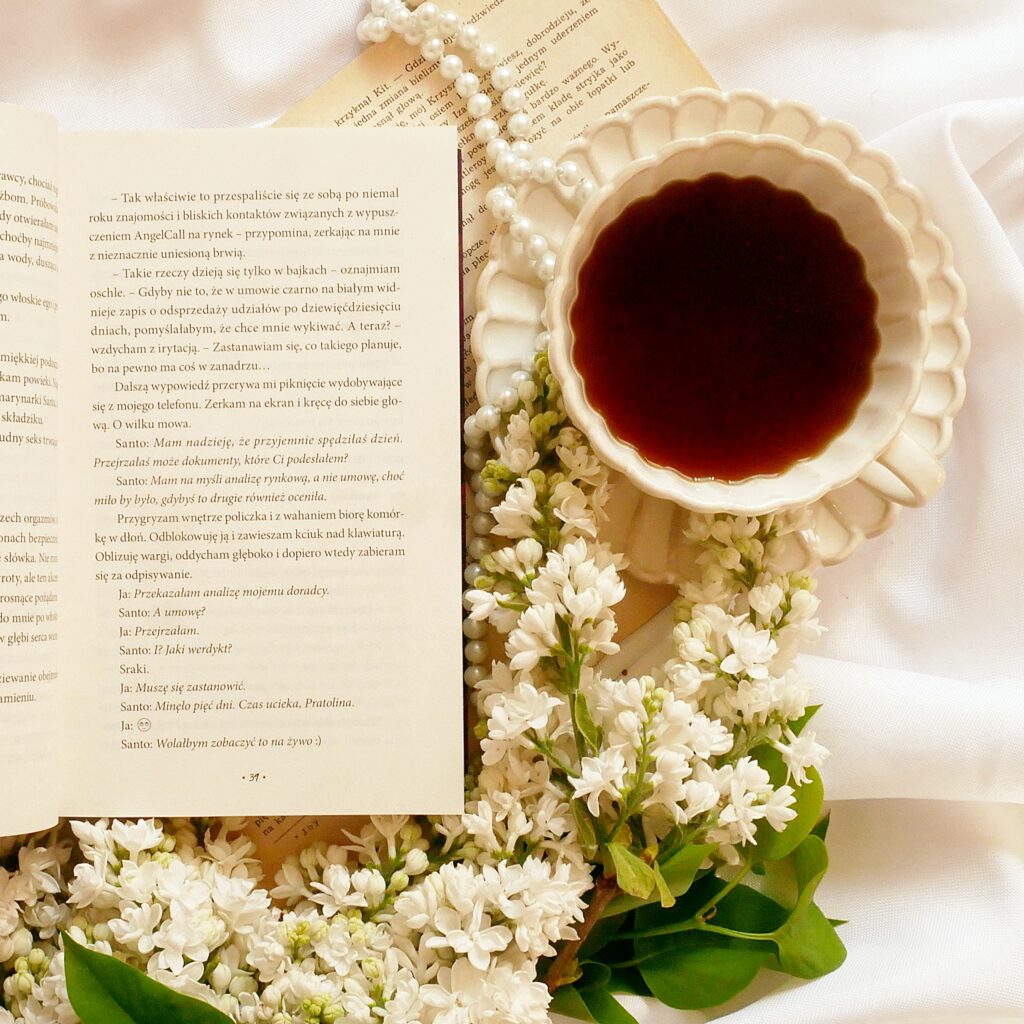 Fragment książki „Lustful” Leny M. Bielskiej Otwarta książka „Lustful” Leny M. Bielskiej na stronie 31, ułożona na białym materiale w otoczeniu filiżanki z czarną kawą, sznura pereł i gałązek bzu.