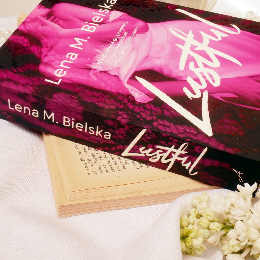 Grzbiet i okładka książki „Lustful” Leny M. Bielskiej Lena M. Bielska „Lustful”. Grzbiet i okładka książki, obok której leżą białe kwiaty bzu.
