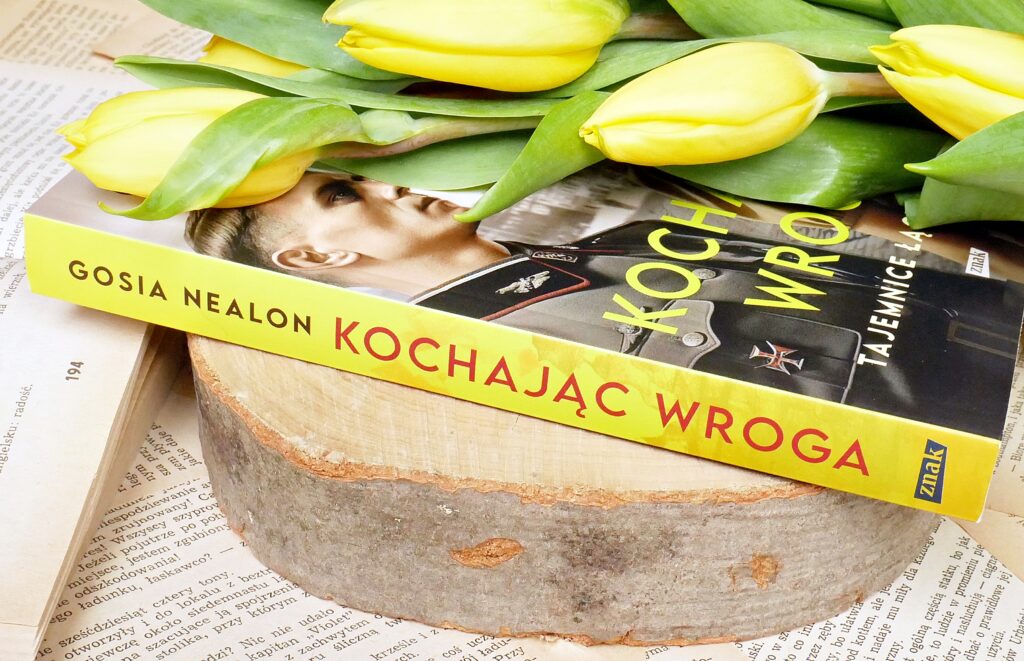 Grzbiet i okładka książki „Kochając wroga” Gosi Nealon Grzbiet książki „Kochając wroga” Gosi Nealon na plastrze drewna, pożółkłych kartkach, starej orwartej książce, z żółtymi tulipanami