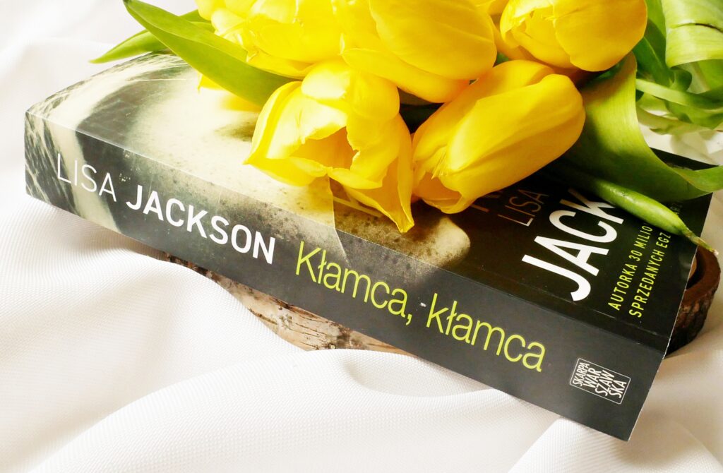 Grzbiet i okładka książki „Kłamca, kłamca” Lisy Jackson Grzbiet książki „Kłamca, kłamca” Lisy Jackson na plastrze drewna z żółtymi tulipanami