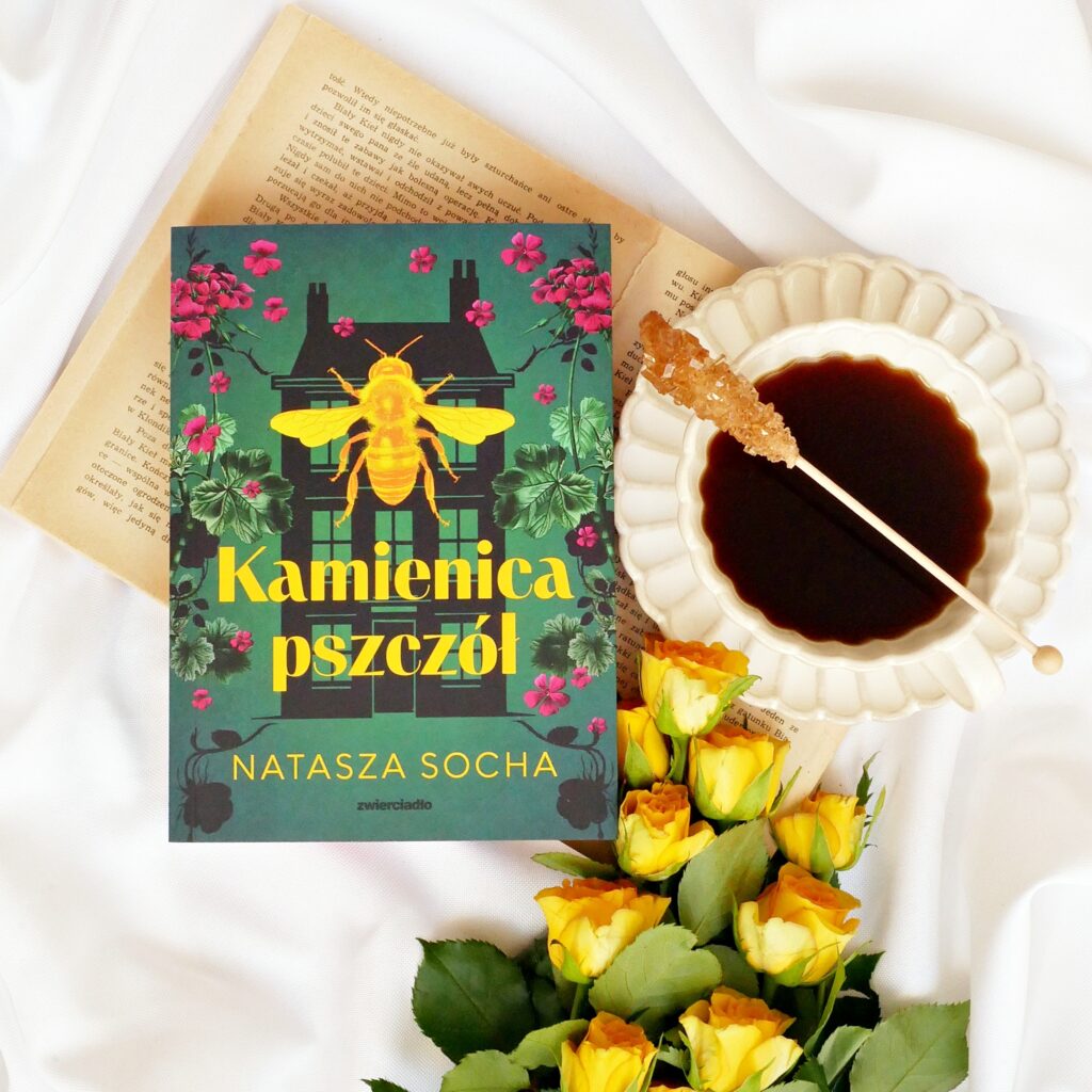 Okładka książki „Kamienica pszczół” Nataszy Sochy. Leży na starej książce, obok filiżanka kawy na spodku i żółte róże.