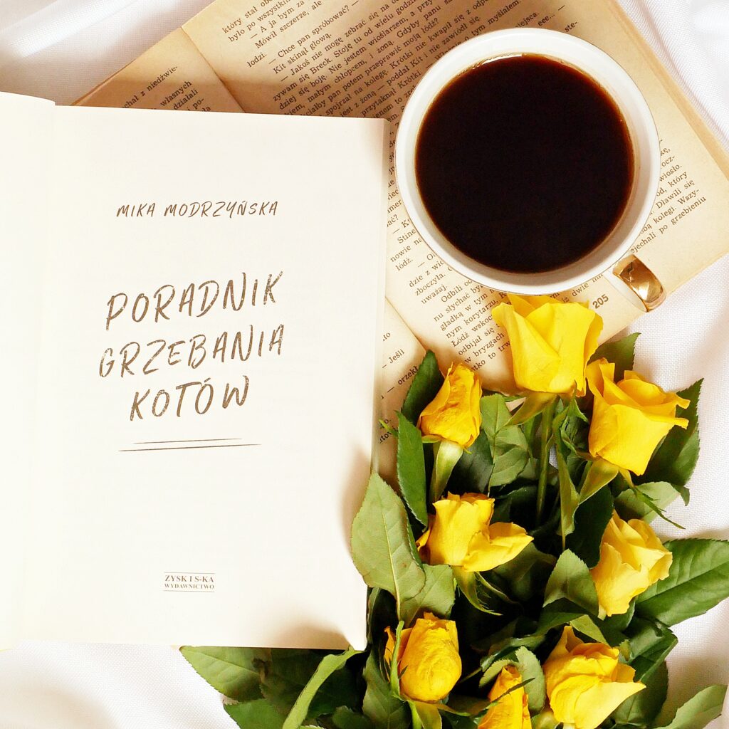 Strona tytułowa książki „Poradnik grzebania kotów” autorstwa Miki Modrzyńskiej Strona tytułowa książki „Poradnik grzebania kotów” autorstwa Miki Modrzyńskiej. Obok leży filiżanka kawy i żółte róże