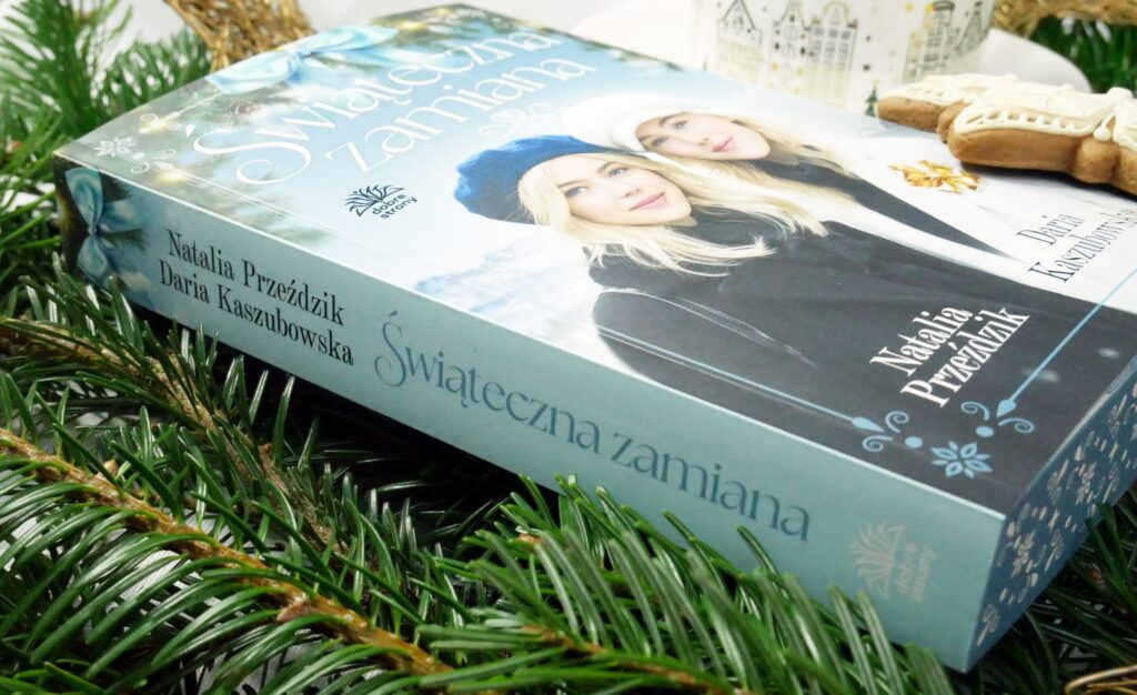 Detale wydania i grzbiet książki„Świąteczna zamiana”. Zbliżenie na błękitny grzbiet książki „Świąteczna zamiana” leżącej wśród gałązek świerku. Na okładce widoczne dwie uśmiechnięte kobiety w zimowych czapkach.