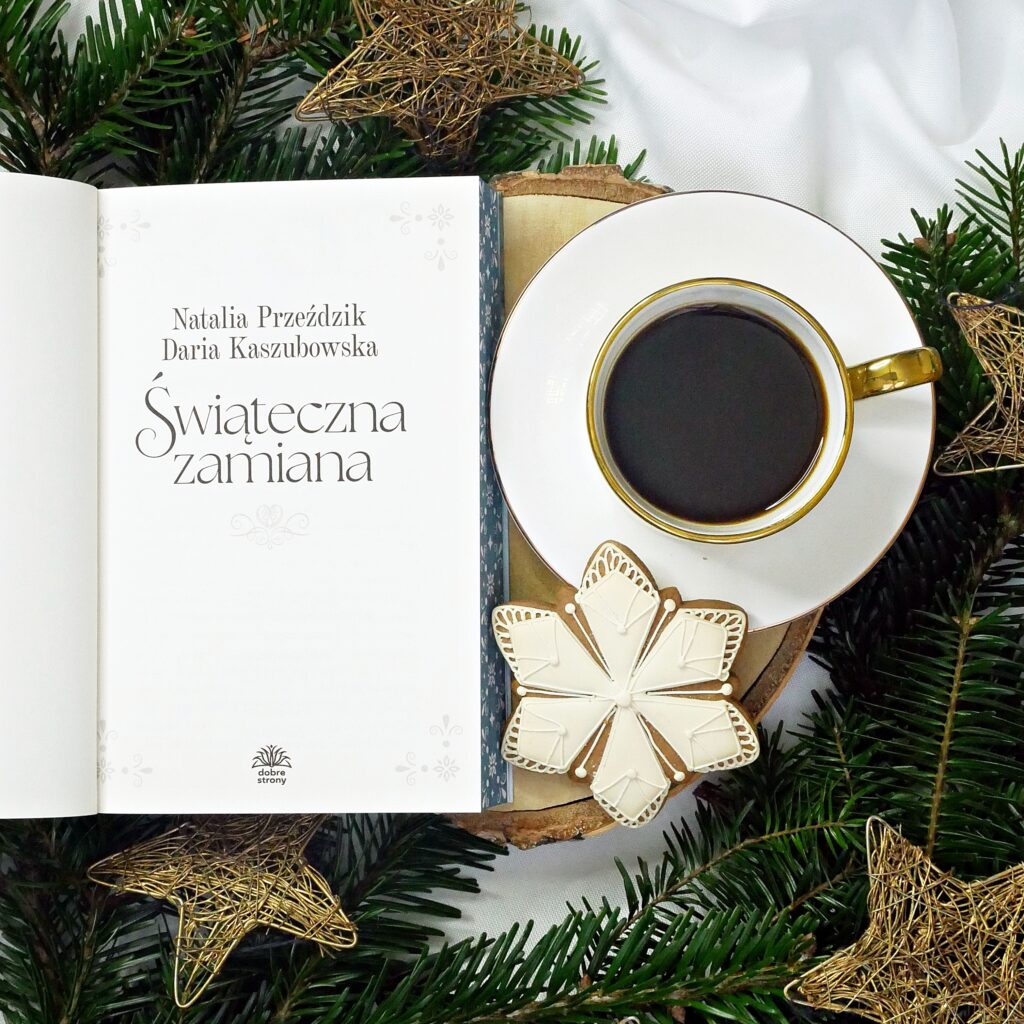 Strona tytułowa książki „Świąteczna zamiana” Otwarta książka na stronie tytułowej z napisem „Świąteczna zamiana” autorstwa Natalii Przeździk i Darii Kaszubowskiej, obok filiżanki czarnej kawy i piernika w kształcie gwiazdy.