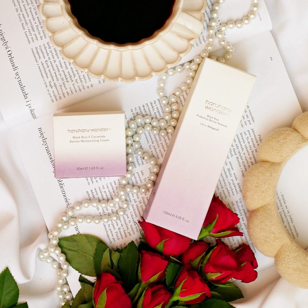 Opakowania esencji i kremu nawilżającego Haruharu Wonder Black Rice leżące na otwartym magazynie. Obok znajduje się filiżanka kawy, opaska, perły i czerwone róże.