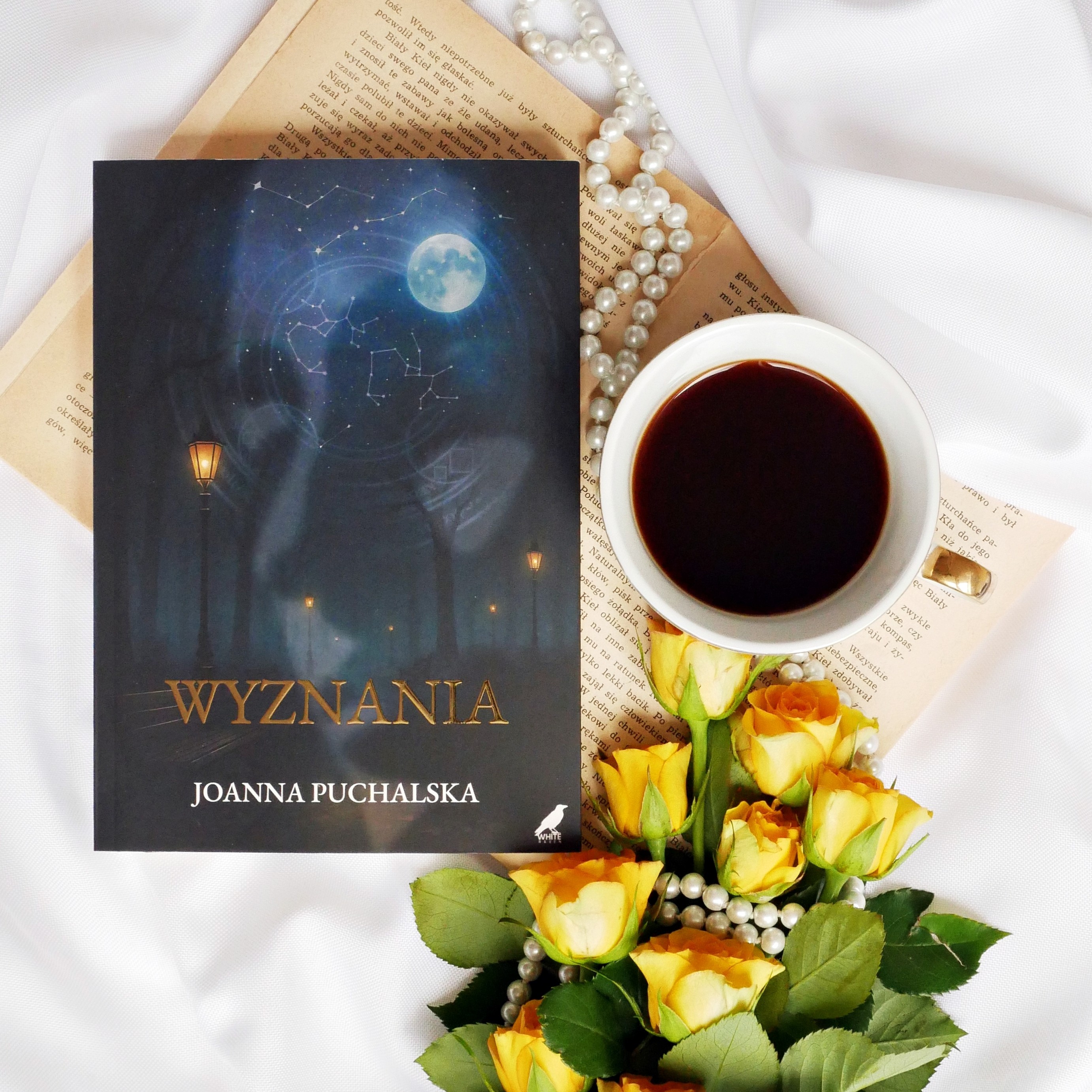 Książka „Wyznania” Joanna Puchalska Okładka książki Joanna Puchalska „Wyznania” z motywem nocnego nieba, gwiazdozbiorów i latarni. Obok leży filiżanka kawy i żółte róże.