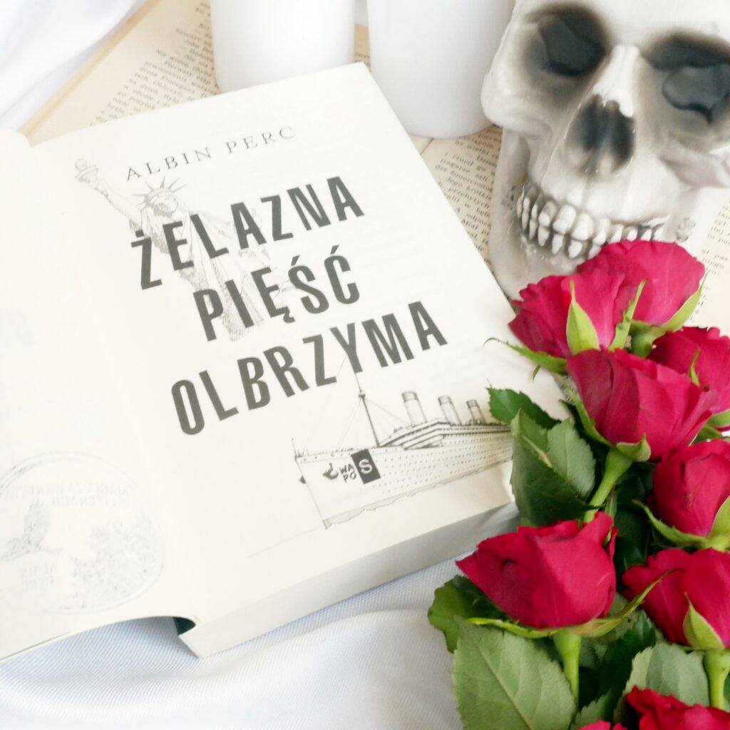 Strona tytułowa książki „Żelazna pięść olbrzyma” autorstwa Albina Perca Strona tytułowa książki „Żelazna pięść olbrzyma” Albina Perca w otoczeniu czerwonych róż i dekoracyjnej czaszki