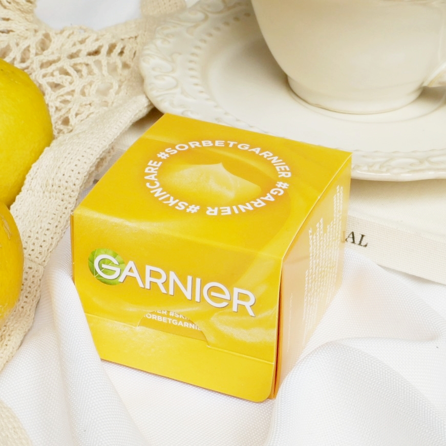 Garnier Vitamin C Fresh & Bright - opakowanie Ozdobny kartonik kremu Garnier Vitamin C Fresh & Bright w letniej, jasnej kompozycji z filiżanką i cytrynami.