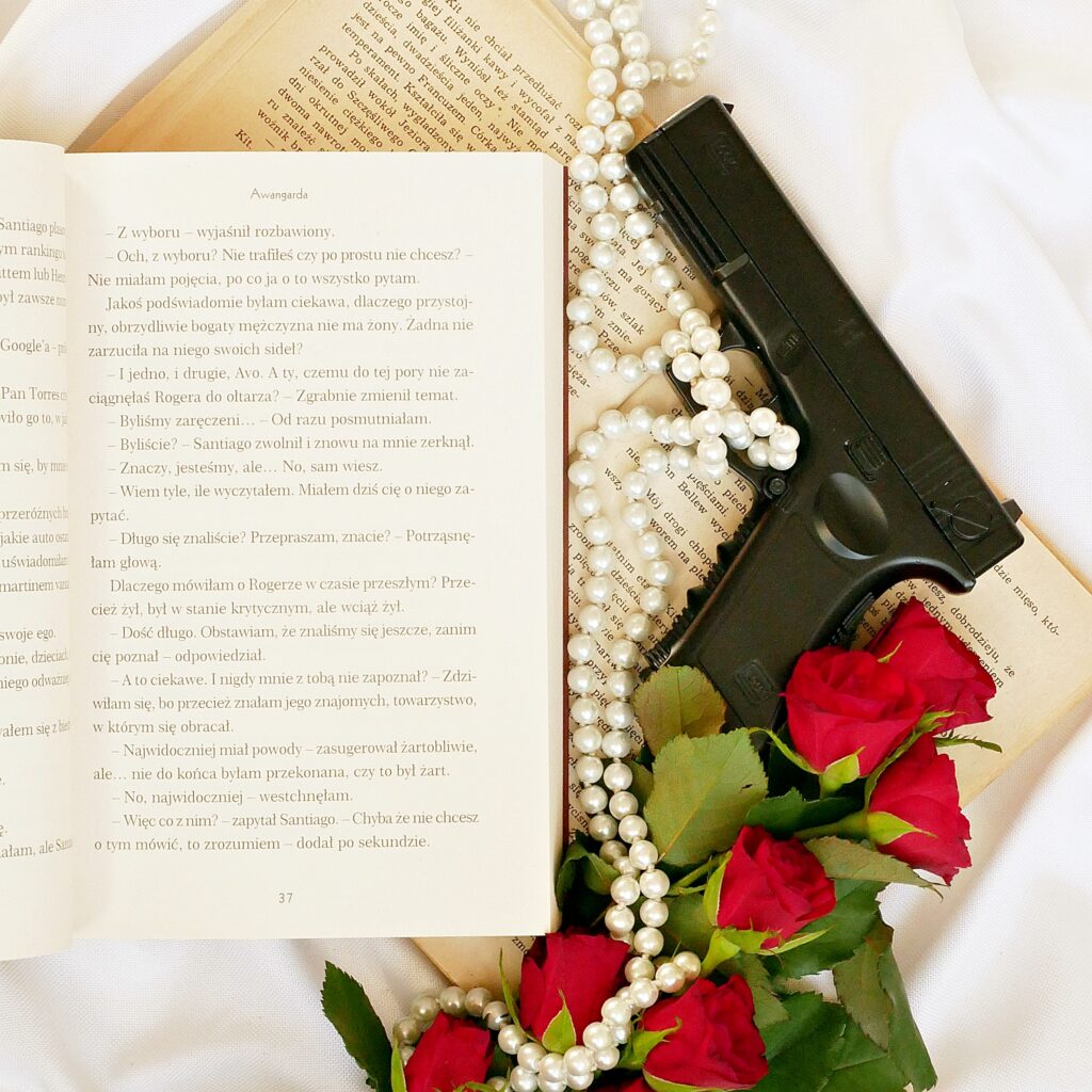 Fragment recenzowanej książki „Awangarda” K.N. Haner Fragment tekstu z książki z dialogami bohaterów. Obok leżą czerwone róże, perły i pistolet.
