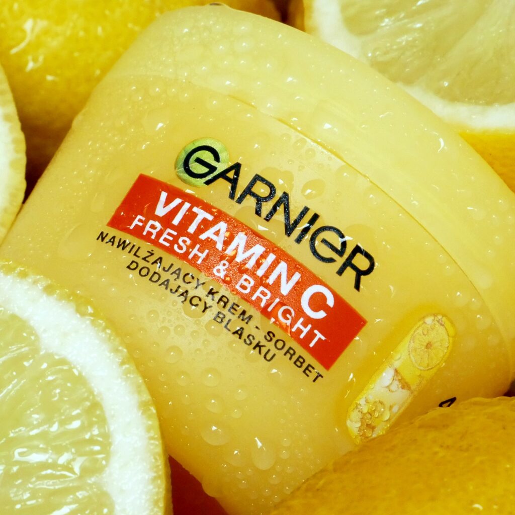 Nawilżający krem Garnier z witaminą C Żółte opakowanie kremu nawilżającego Garnier Vitamin C Fresh & Bright pokryte kroplami wody, z połówkami świeżych cytryn w tle.