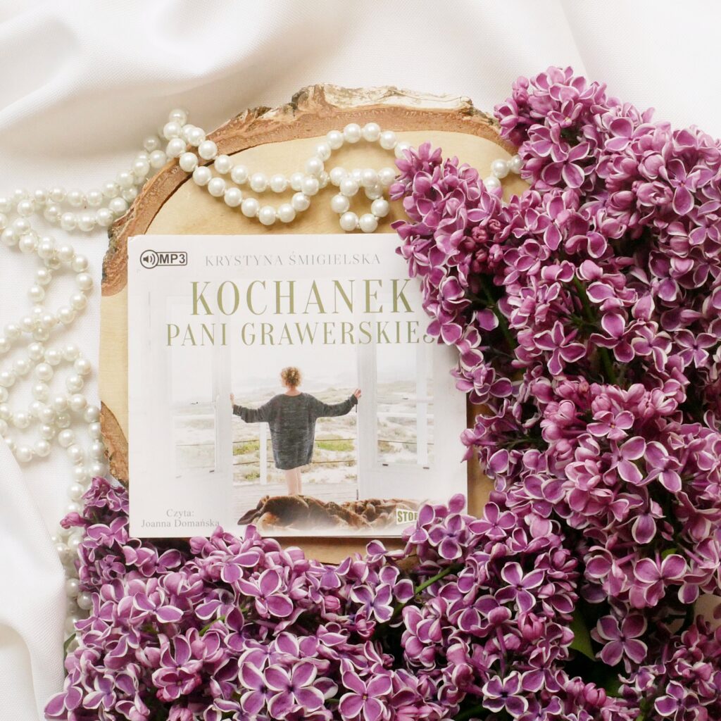 Krystyna Śmigielska „Kochanek pani Grawerskiej” - recenzja Płyta z audiobookiem „Kochanek pani Grawerskiej” leżąca na drewnie, w otoczeniu pereł i fioletowego bzu.