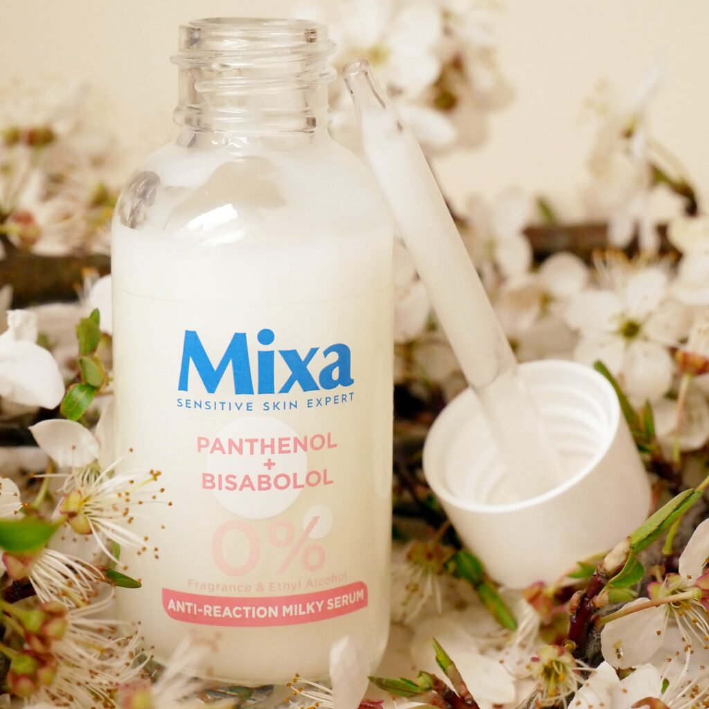 Serum Mixa Anti Reaction z pipetą Otwarte opakowanie mlecznego serum kojącego Mixa Anti Reaction z widoczną obok pipetą na tle kwitnących kwiatów jabłoni.