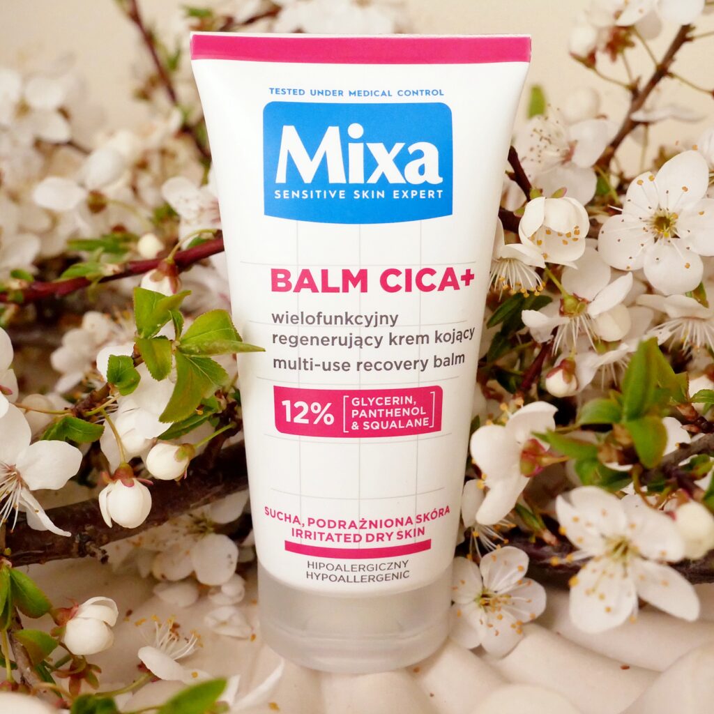 Krem regenerujący i kojący Mixa Balm Cica+ Opakowanie wielofunkcyjnego kremu regenerującego i kojącego Mixa Balm Cica+ wśród kwitnących kwiatów jabłoni.