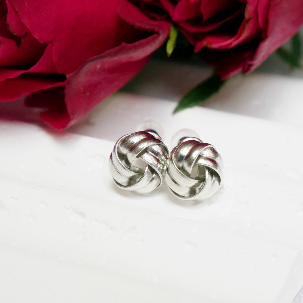 Srebrne kolczyki Silver Nido – elegancki pomysł na prezent na Dzień Matki Srebrne kolczyki sztyfty Silver Nido o splocie węzła, ułożone na białym tle w towarzystwie czerwonych róż. Elegancka biżuteria na prezent z okazji Dnia Matki.