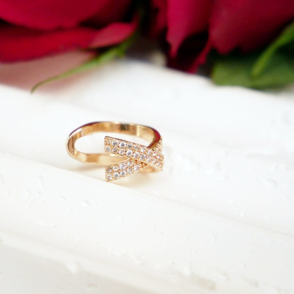 Złota nausznica Diamond X od Gerlinde – elegancki prezent na Dzień Matki Złota nausznica Diamond X marki Gerlinde, ozdobiona skrzyżowanymi pasmami cyrkonii. Biżuteria wyeksponowana na białej, schodkowej podstawce w otoczeniu czerwonych róż.