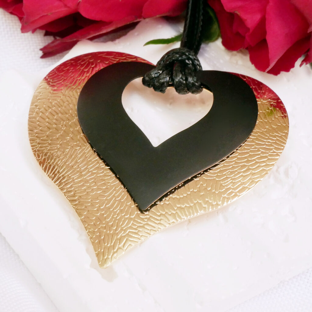 Złoto-czarny naszyjnik Heartline – elegancki prezent na Dzień Matki Zbliżenie na elegancki, złoto-czarny naszyjnik Heartline w kształcie podwójnego serca. Biżuteria leży na białej podstawce w otoczeniu czerwonych róż jako propozycja na luksusowy prezent na Dzień Matki.