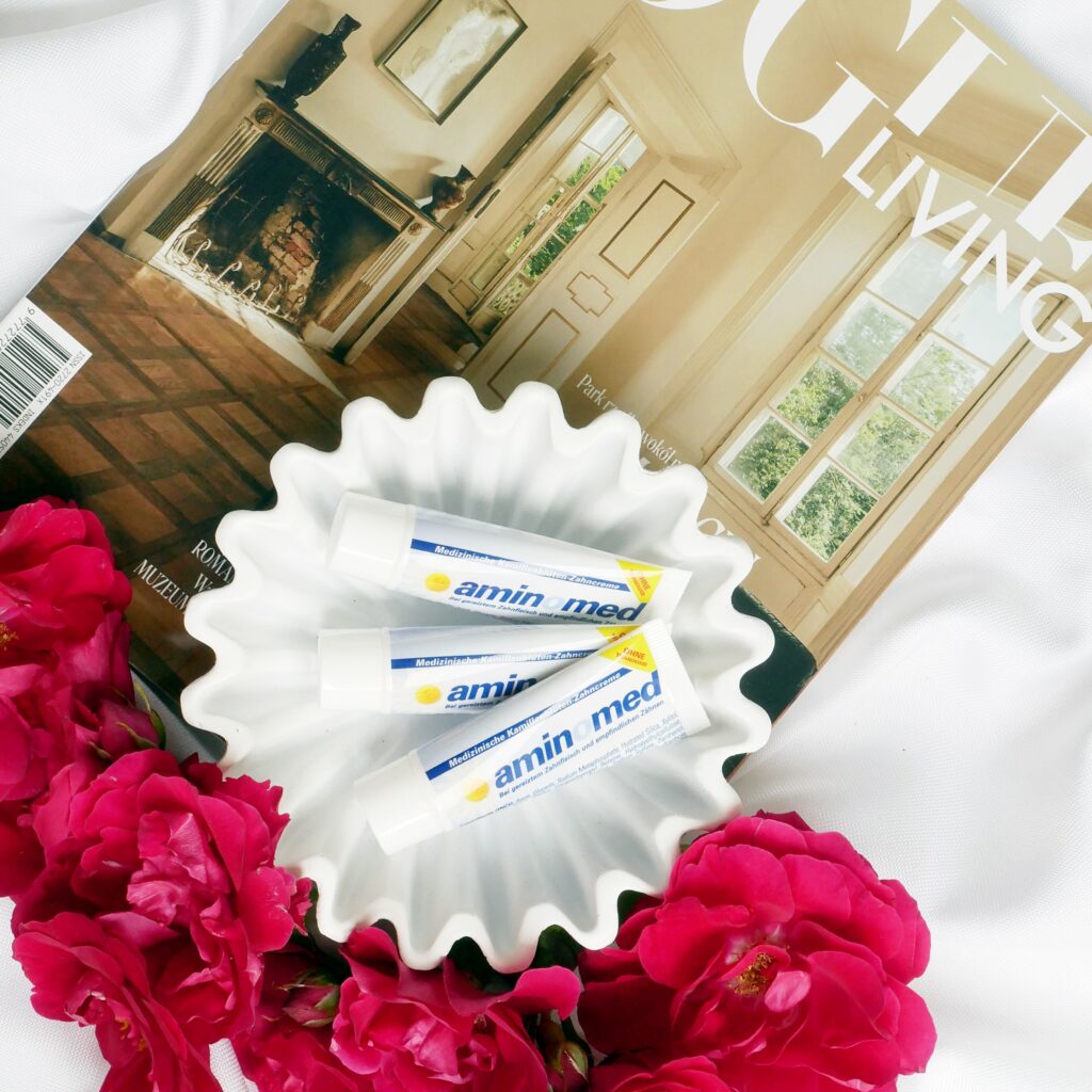 Trzy tubki łagodnej pasty Aminomed na wrażliwe zęby ułożone w białej miseczce na tle magazynu Vogue Living w towarzystwie czerwonych róż.
