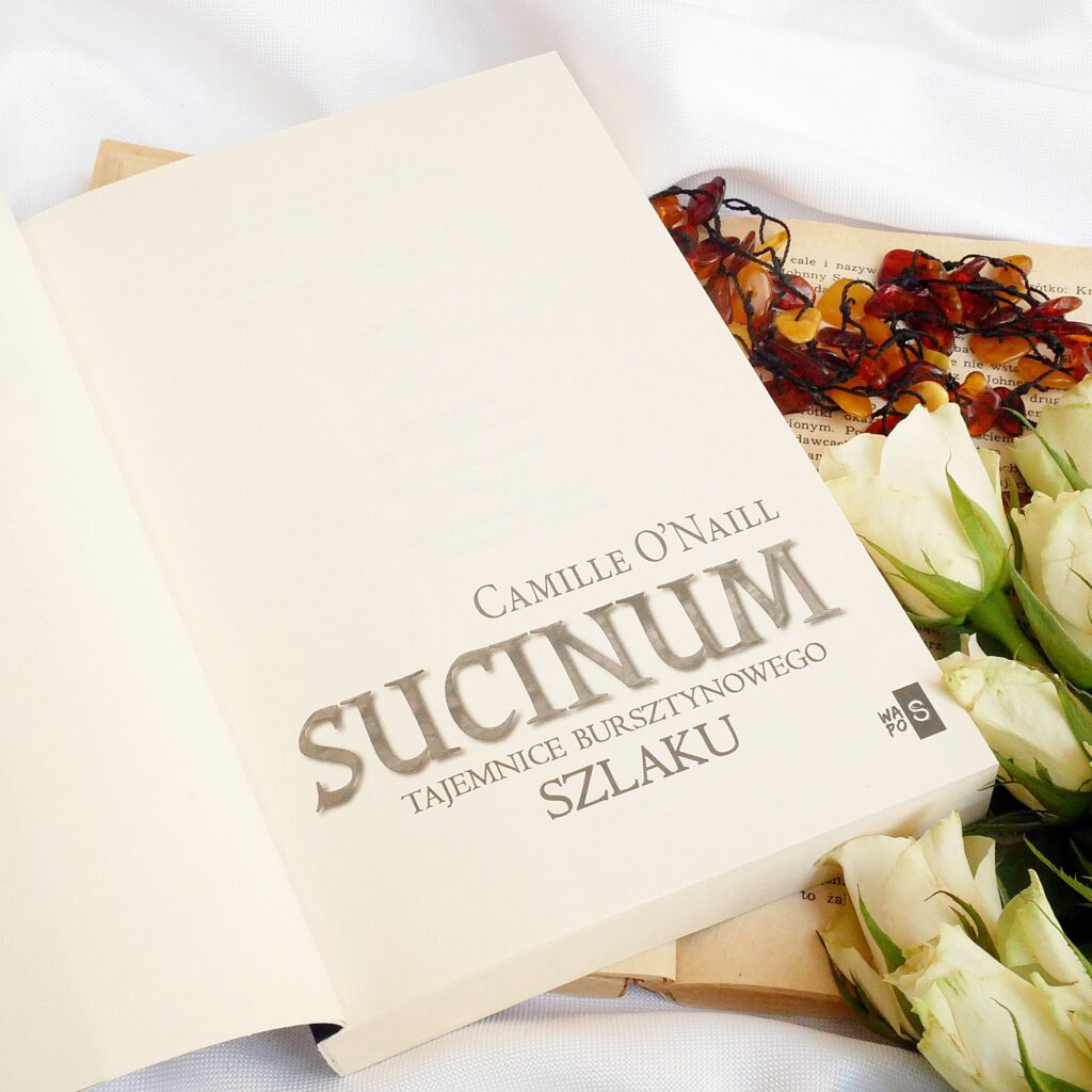 Strona tytułowa książki Camille O’Neill „Sucinum. Tajemnice bursztynowego szlaku” w klimatycznej aranżacji z białymi różami i bursztynowym naszyjnikiem.