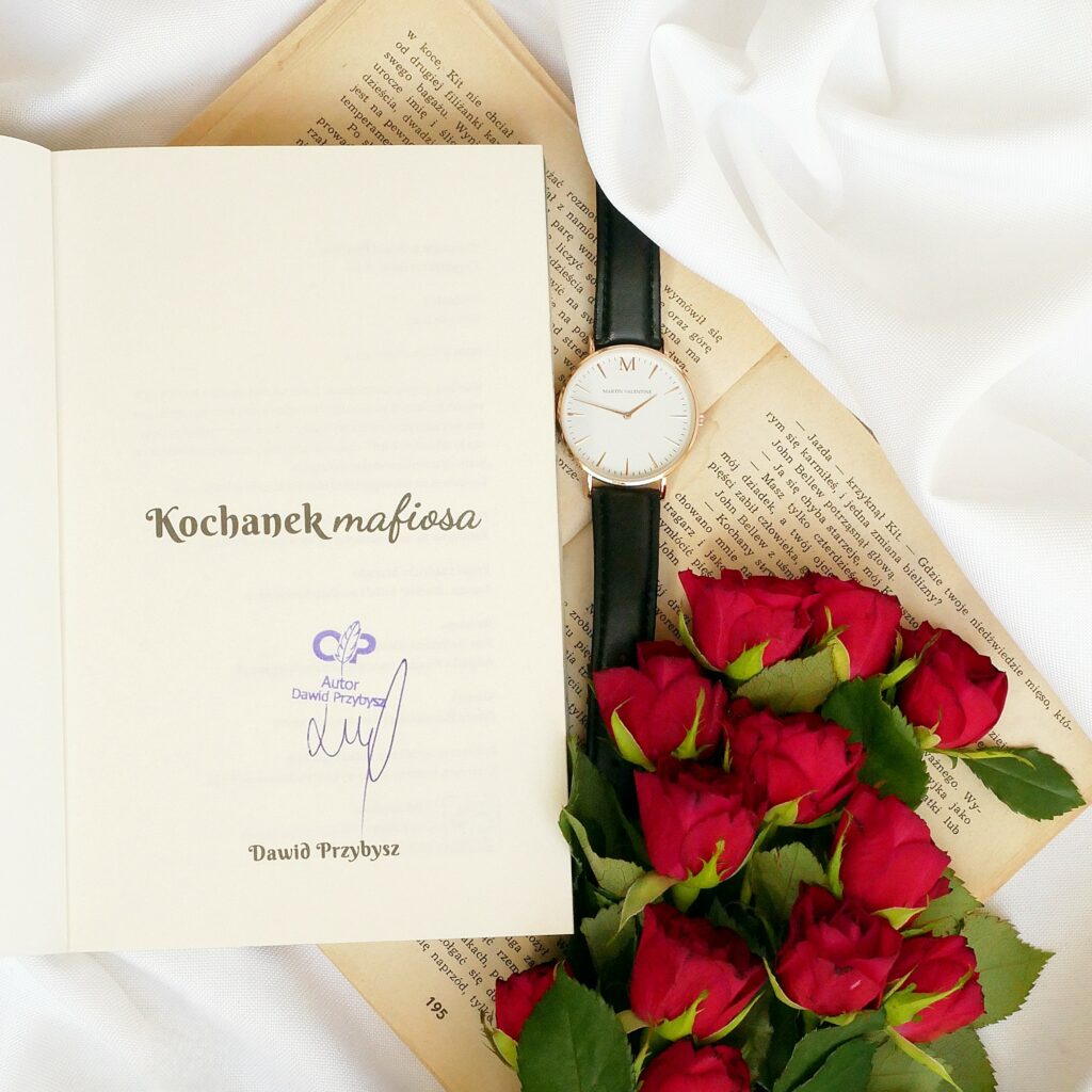 Strona tytułowa książki „Kochanek mafiosa” z autografem i fioletową pieczątką autora Dawida Przybysza. Kompozycja z zegarkiem i czerwonymi różami.