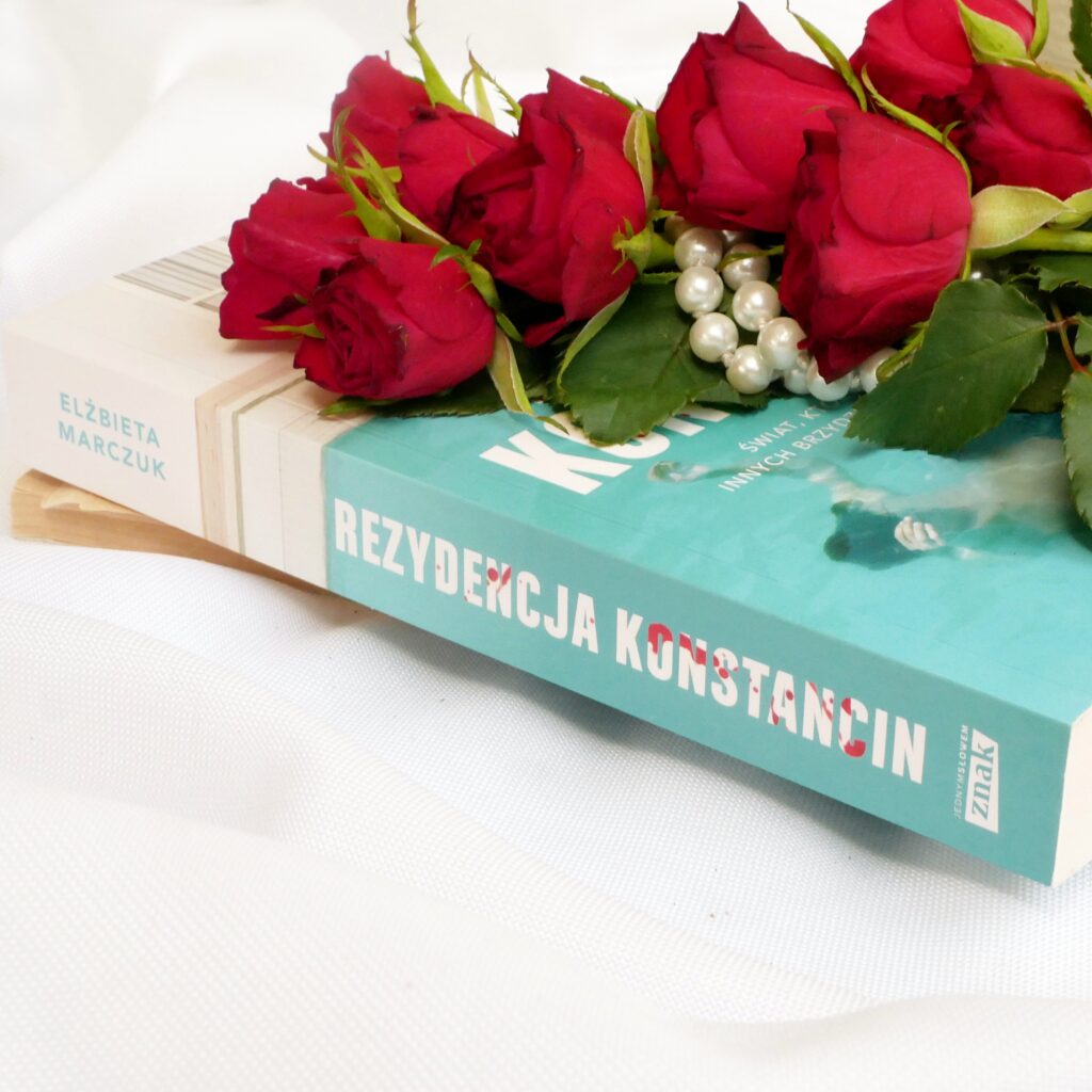 Zbliżenie na grzbiet książki Elżbiety Marczuk „Rezydencja Konstancin” Zbliżenie na grzbiet książki Elżbiety Marczuk „Rezydencja Konstancin” w otoczeniu czerwonych róż i sznura pereł,