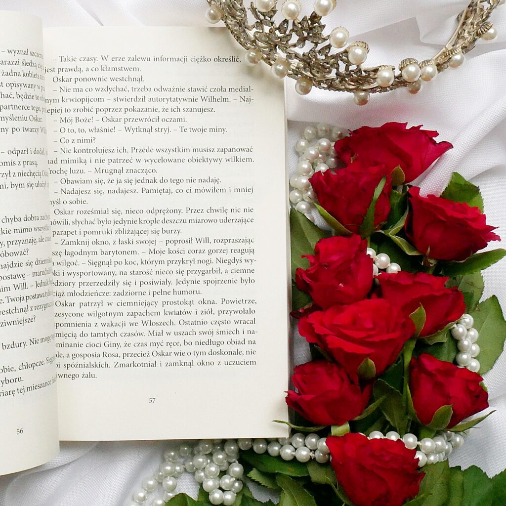 Otwarta książka Katarzyny Redmerskiej „Jak (nie) zostać królową”na stronie 57 w otoczeniu czerwonych róż, pereł i korony.