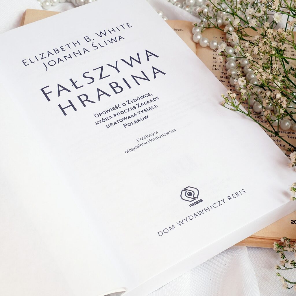 Strona tytułowa książki „Fałszywa hrabina” leżąca na białym materiale i starej książce w otoczeniu pereł i gipsówki.