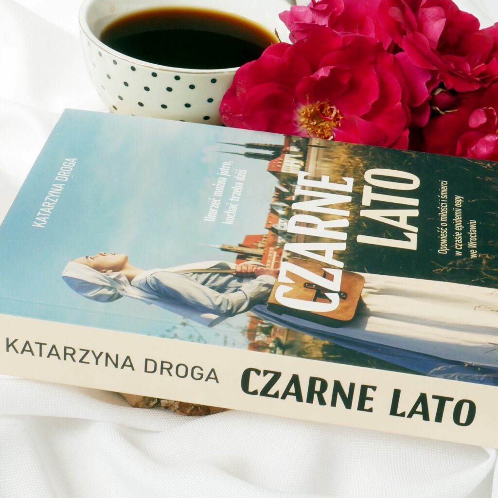 Grzbieti książki Katarzyny Drogi „Czarne lato” Grzbieti książki Katarzyny Drogi „Czarne lato” w otoczeniu czerwonych róż i filiżanki czarnej kawy.