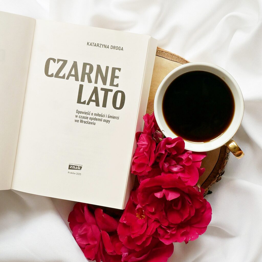 Strona tytułowa książki Katarzyny Drogi „Czarne lato” Strona tytułowa książki Katarzyny Drogi „Czarne lato” w otoczeniu czerwonych róż i filiżanki czarnej kawy.