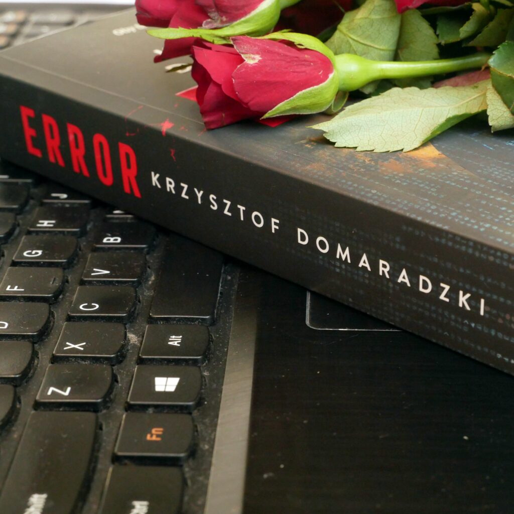 Grzbiet i okładka książki „Error” Krzysztofa Domaradzkiego Krzysztof Domaradzki „Error”. Grzbiet i okładka książki z leżącymi na niej czerwonymi różami, ułożona na klawiaturze czarnego laptopa.