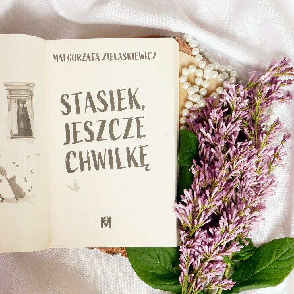 Strona tytułowa książki „Stasiek jeszcze chwilkę” Małgorzaty Zielaskiewicz. Strona tytułowa książki „Stasiek jeszcze chwilkę” Małgorzaty Zielaskiewicz. Książka ułożona w estetycznej kompozycji na jasnym tle w otoczeniu pereł i gałązek fioletowego bzu.