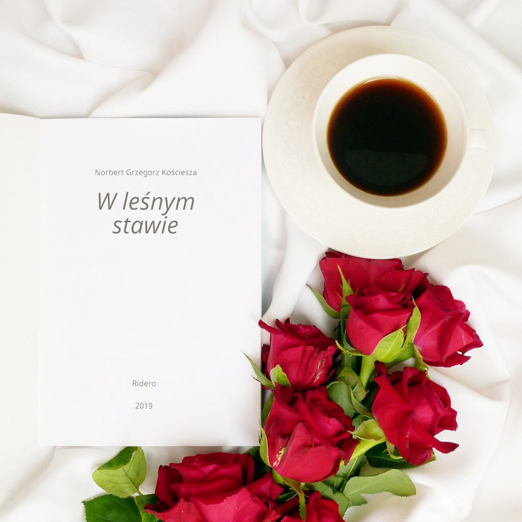 Strona tytułowa książki Norberta Grzegorza Kościeszy „W leśnym stawie” Strona tytułowa książki z napisem Norbert Grzegorz Kościesza „W leśnym stawie” na białym materiale w otoczeniu czerwonych róż i filiżanki kawy.