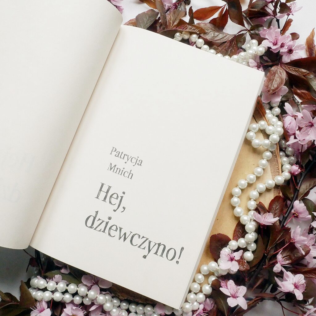 Patrycja Mnich „Hej, dziewczyno!”. Strona tytułowa książki ułożona na jasnym tle na plastrze drewna, w otoczeniu pereł i drobnych, różowych kwiatów wiśni.