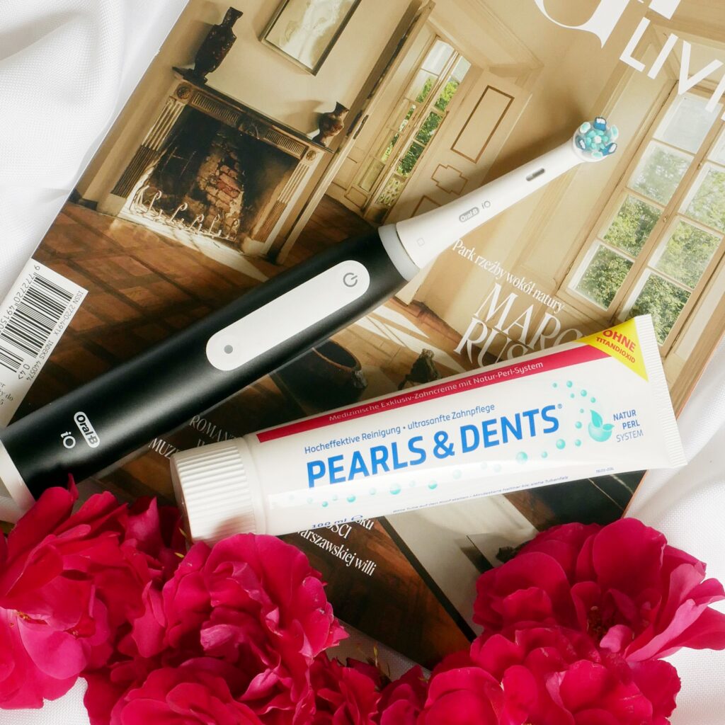Pasta do zębów Pearls & Dents oraz czarna szczoteczka elektryczna Oral-B leżące na tle magazynu Vogue Living w towarzystwie czerwonych róż.
