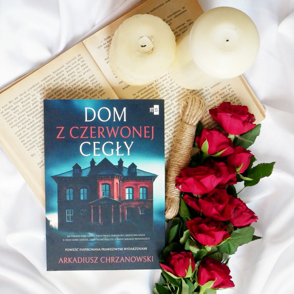 Arkadiusz Chrzanowski „Dom z czerwonej cegły” - recenzja Okładka książki Arkadiusza Chrzanowskiego „Dom z czerwonej cegły” na tle białego, pomarszczonego materiału. Dopracowany kadr z czerwonymi różami, świecami i starymi, pożółkłymi stronami.