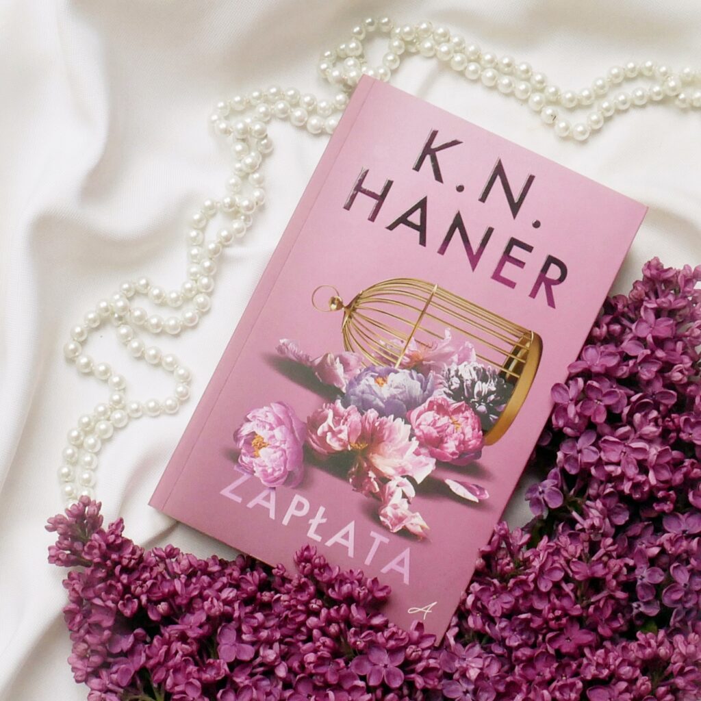 K.N. Haner „Zapłata” - recenzja Okładka książki K.N. Haner „Zapłata” ze złotą klatką, leżąca na białym materiale obok fioletowych bzów i pereł.