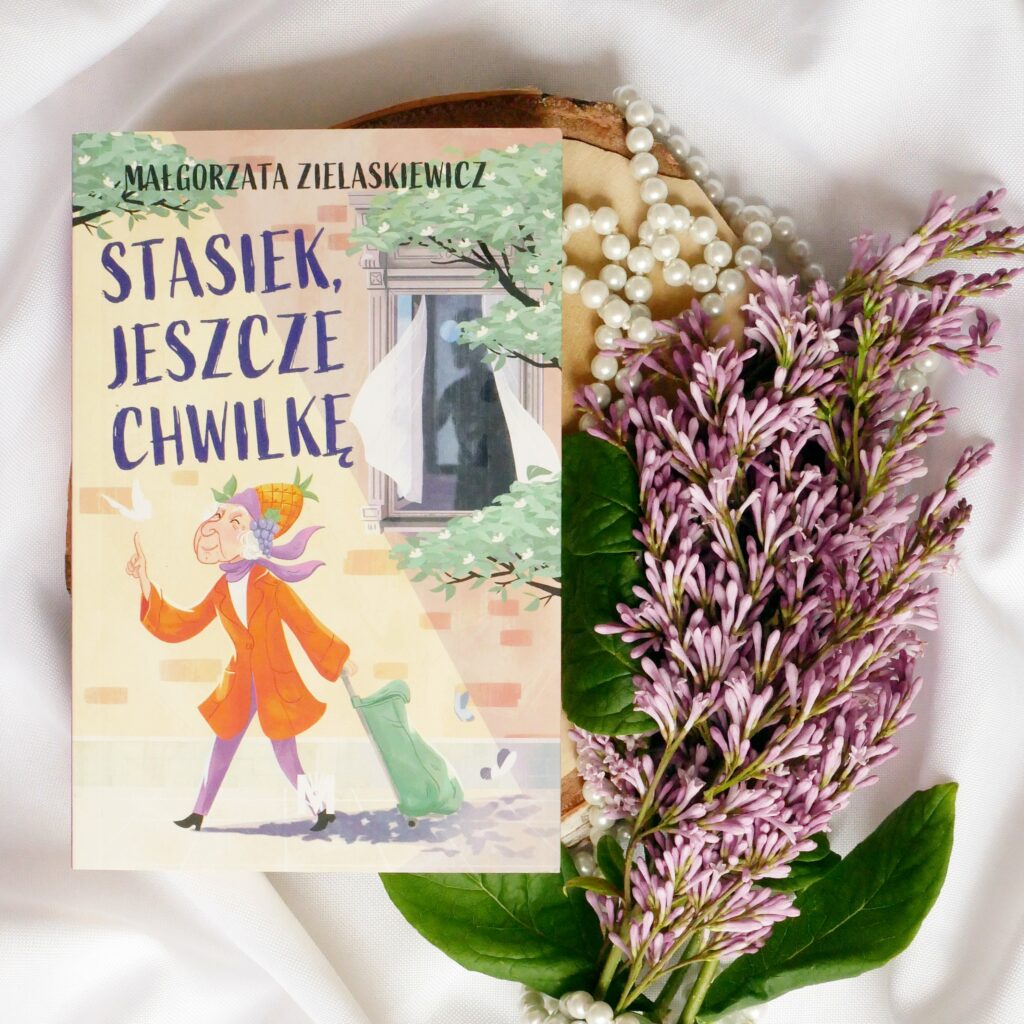 Małgorzata Zielaskiewicz „Stasiek jeszcze chwilkę” - recenzja Okładka książki „Stasiek jeszcze chwilkę” Małgorzaty Zielaskiewicz. Książka ułożona w estetycznej kompozycji na jasnym tle w otoczeniu pereł i gałązek fioletowego bzu.