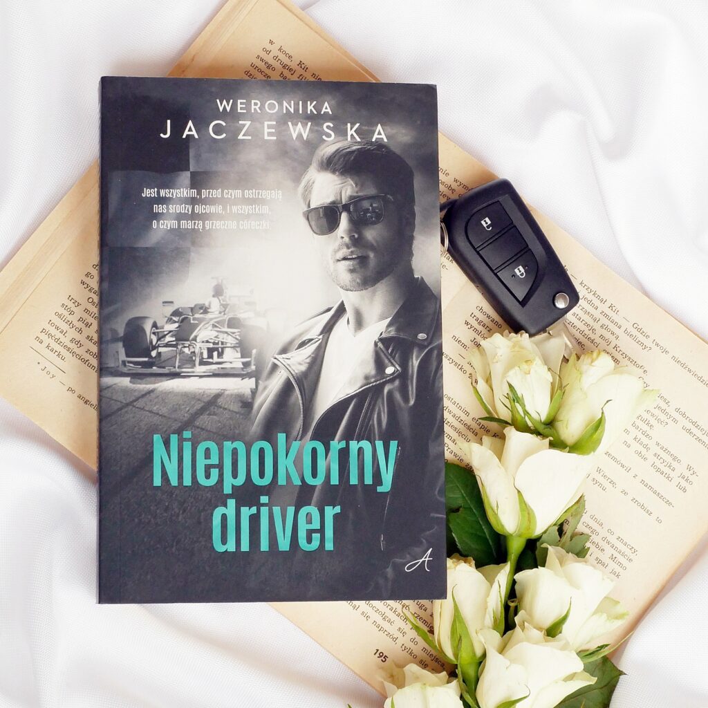 Okładka książki Weronika Jaczewska „Niepokorny driver” w otoczeniu białych róż i kluczyków samochodowych.