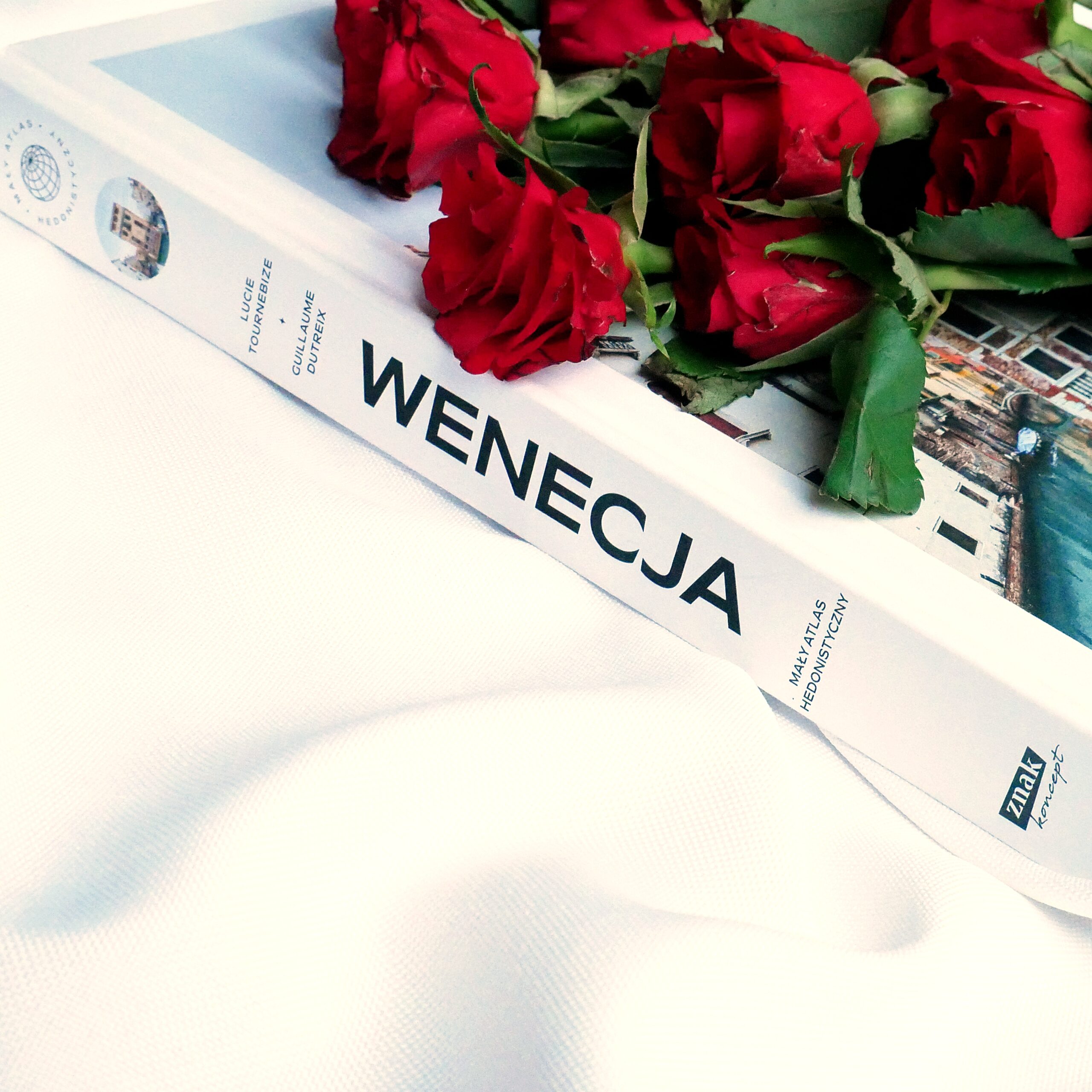 Zbliżenie na gruby grzbiet albumu „Wenecja. Mały atlas hedonistyczny” w otoczeniu świeżych, czerwonych róż na tle białego materiału.