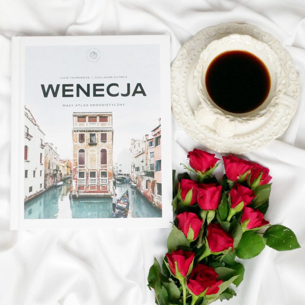 Okładka albumu „Wenecja. Mały atlas hedonistyczny” z widokiem na kanał i kamienicę w kompozycji z filiżanką czarnej kawy i bukietem świeżych, czerwonych róż.