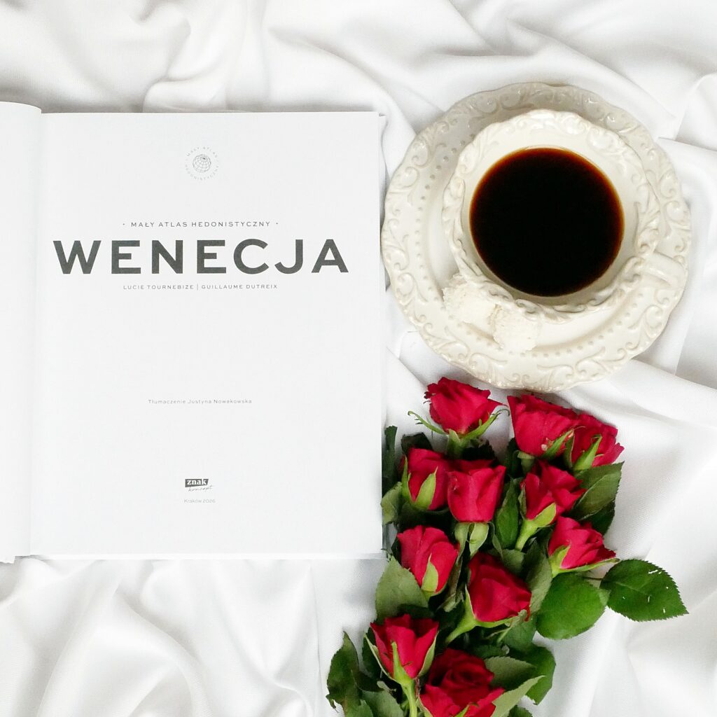 Strona tytułowa książki „Wenecja. Mały atlas hedonistyczny” autorstwa Lucie Tournebize i Guillaume Dutreix, ułożona obok filiżanki kawy oraz świeżych, czerwonych róż.