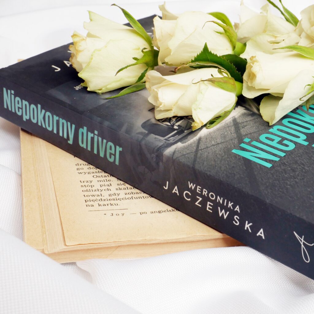Grzbiet i okładka książki „Niepokorny driver” Weroniki Jaczewskiej, na której ułożono świeże, białe róże.