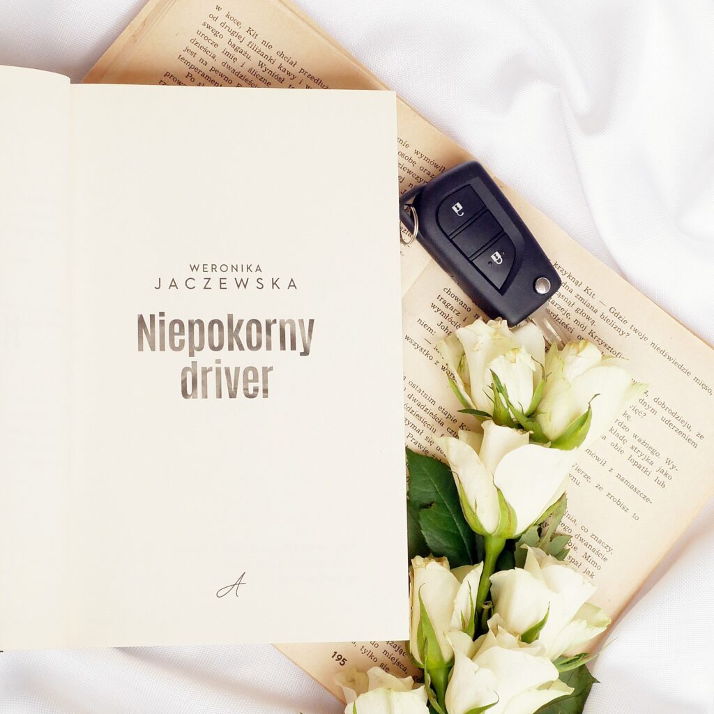 Strona tytułowa książki „Niepokorny driver” Weroniki Jaczewskiej w estetycznej aranżacji typu flatlay z białymi różami i kluczykami do samochodu.