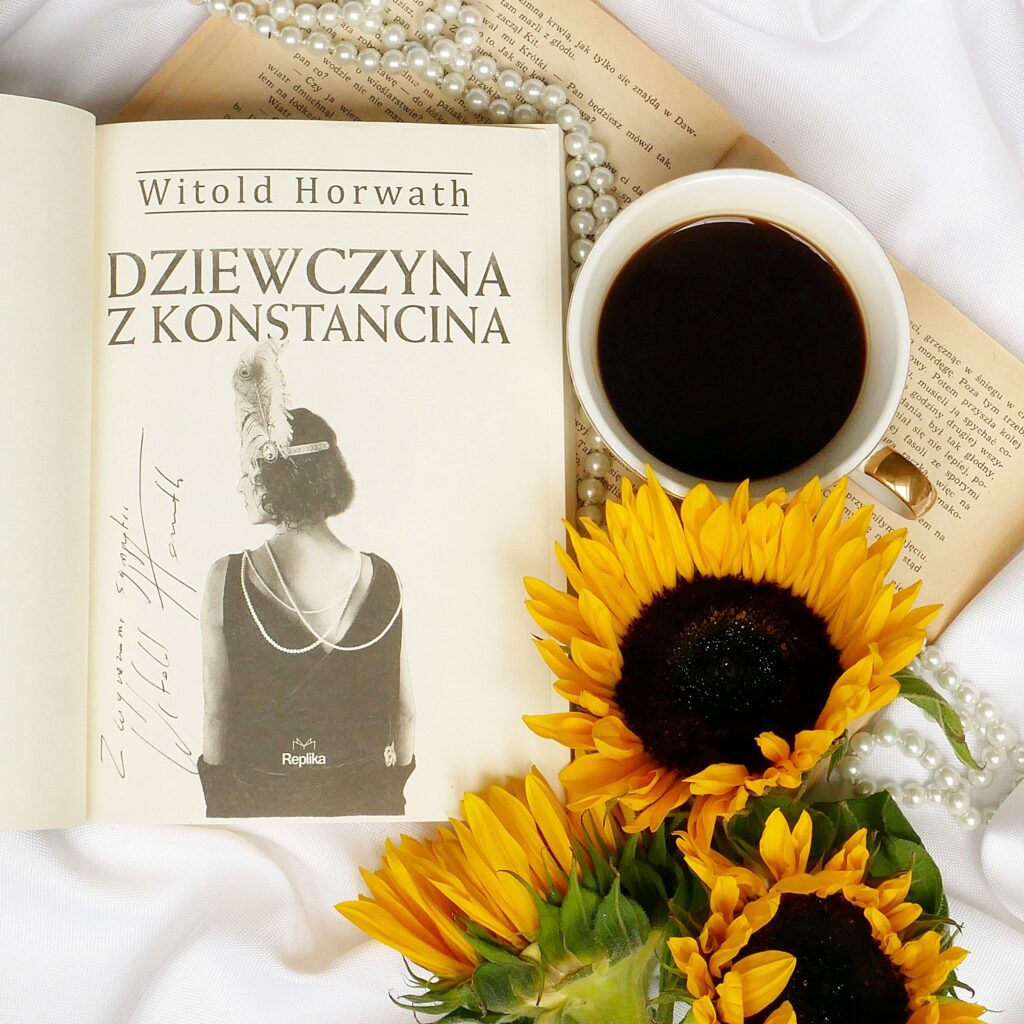 Strona tytułowa książki „Dziewczyna z Konstancina” z odręcznym autografemd autora Witolda Horwatha. Książka w otoczeniu słoneczników, z perłami i filiżanką kawy. 
