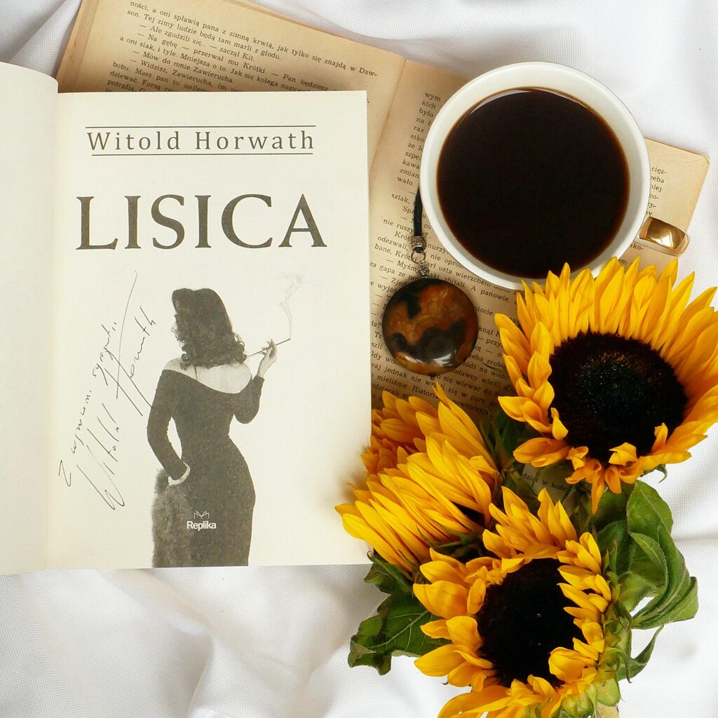 Strona tytułowa książki „Lisica” z odręcznym autografemd autora Witolda Horwatha. Książka w otoczeniu słoneczników, z naszyjnikiem i filiżanką kawy. 